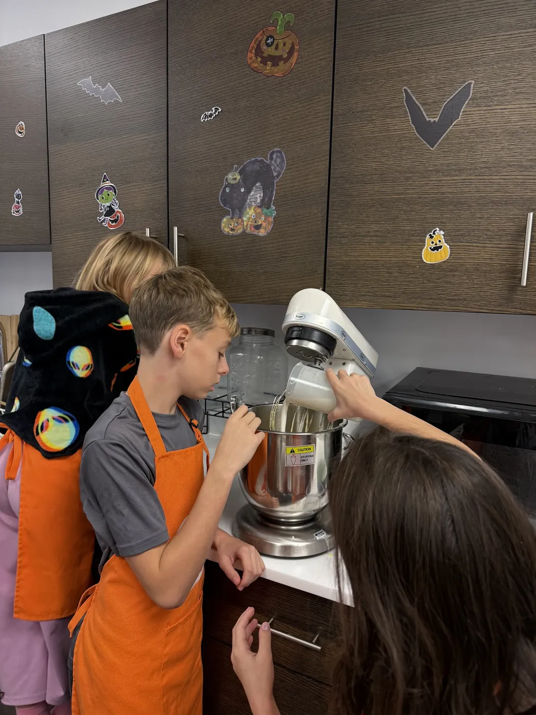 Children in orange aprons add ingredients to a stand mixer during a Cookiddoo cooking class