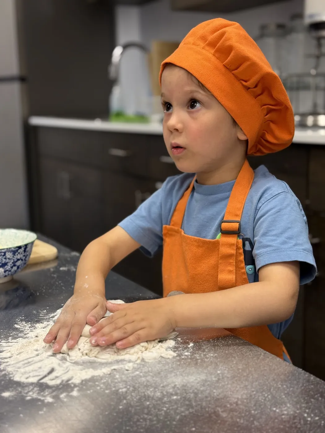 Boy in an orange Cookiddoo chef hat kneading dough on a floured counter at a winter cooking class