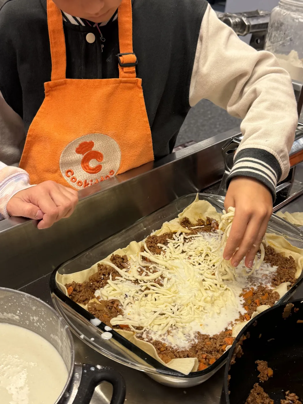 A child in an apron spreads grated cheese over a tray of lasagna