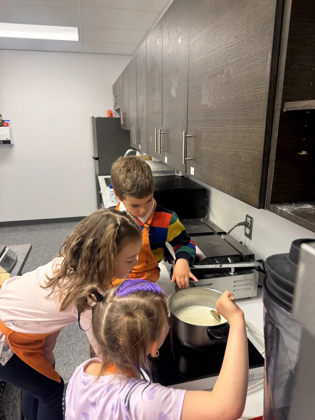 Three children in orange aprons stir a pot of white batter on an induction cooktop during a Cookiddoo cooking class