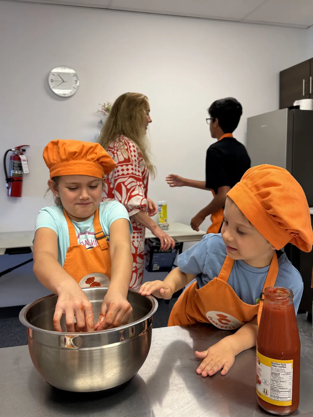 Girl crushing tomatoes in a bowl at a Cookiddoo class while an instructor watches