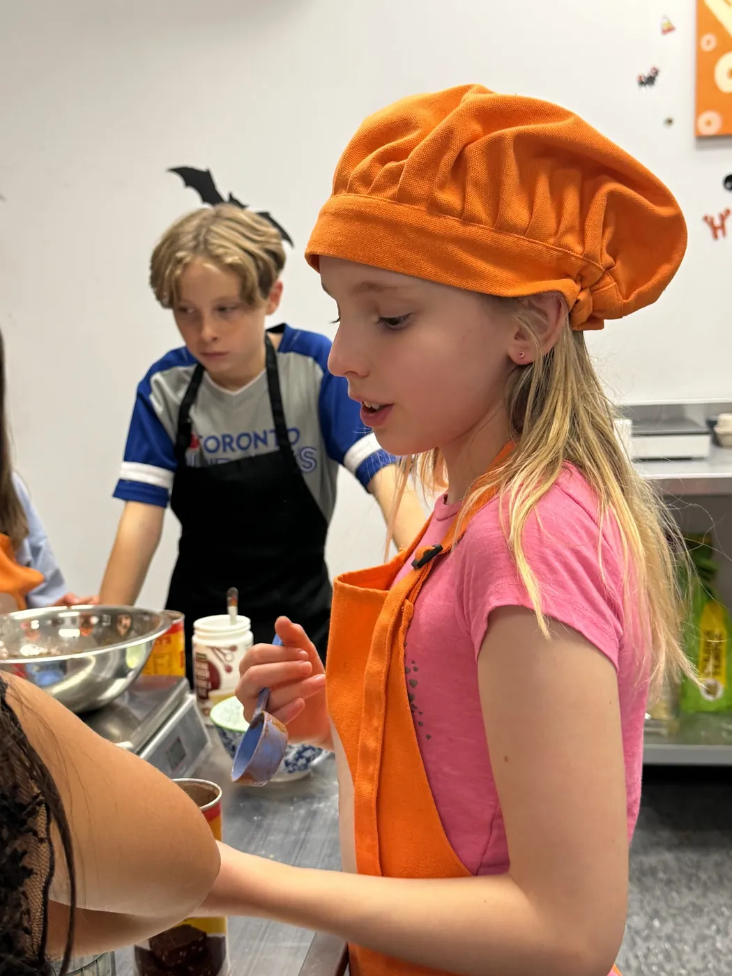 Girl in an orange Cookiddoo chef hat pouring ingredients from a bowl during a kids cooking class