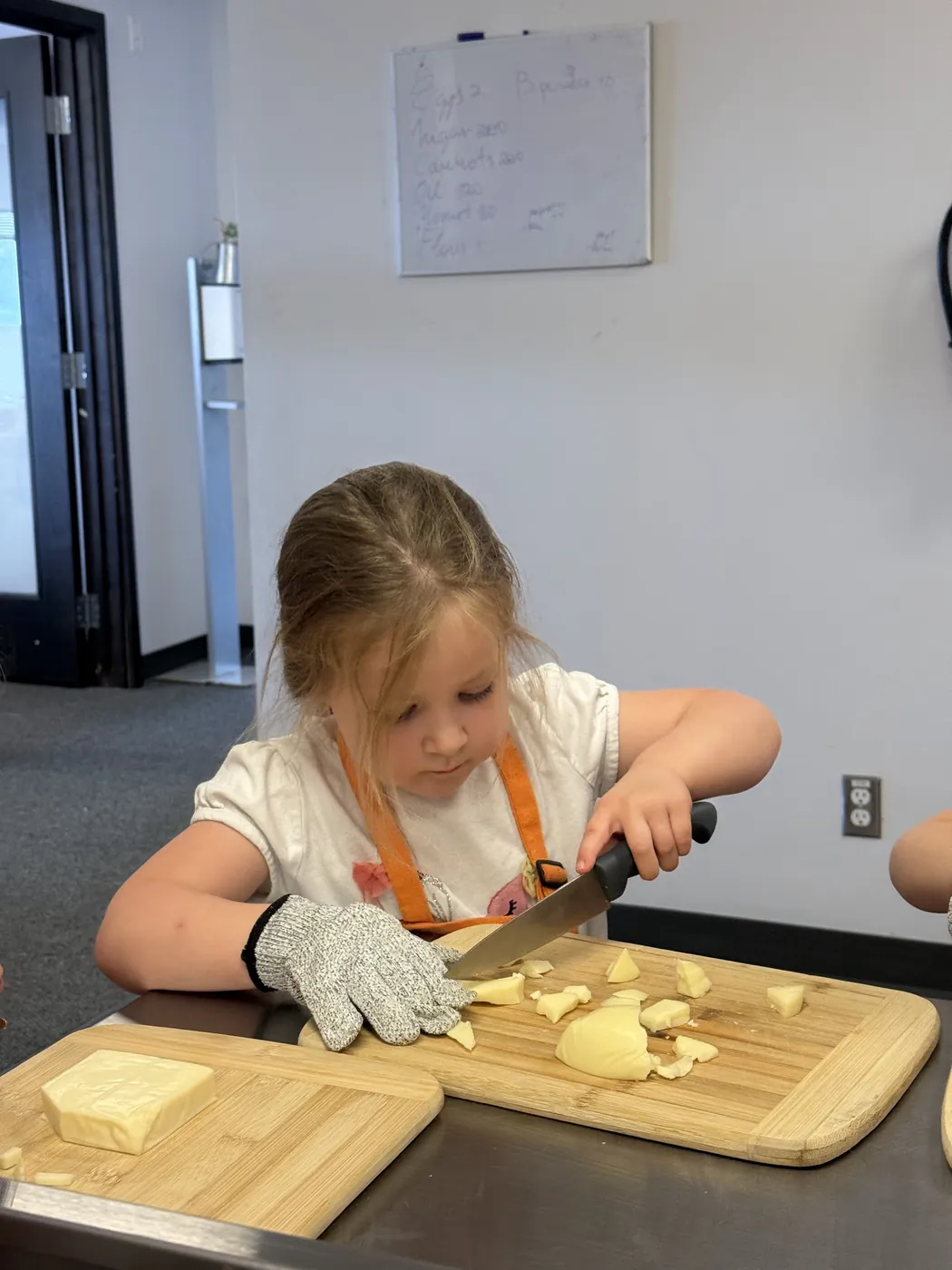 Young girl cutting cheese on a wooden board with a cut-resistant glove at her first Cookiddoo cooking class