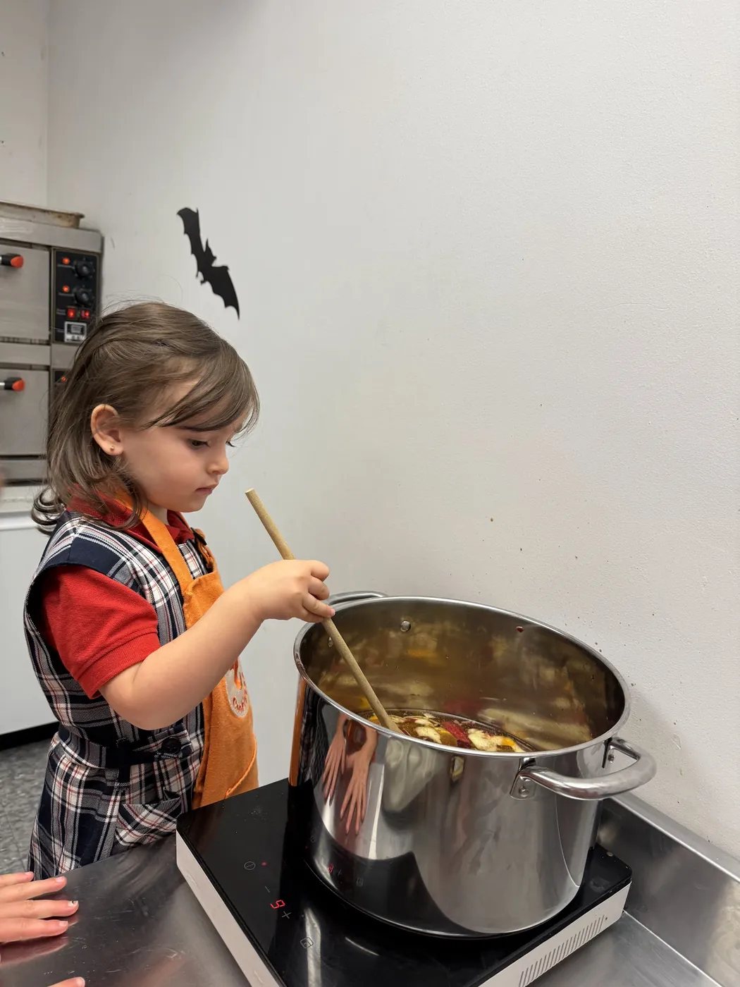 Young girl in a Cookiddoo apron stirring a large pot on the stove during a cooking class
