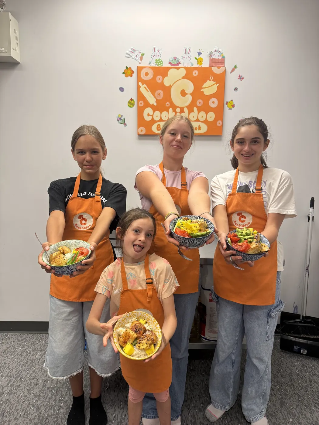 Four girls in Cookiddoo aprons holding up the bowls they cooked at a kids cooking class in Ottawa
