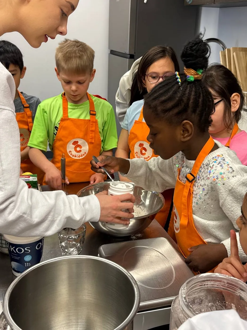 Cookiddoo instructor adding flour to a mixing bowl while kids in orange aprons watch during a cooking lesson