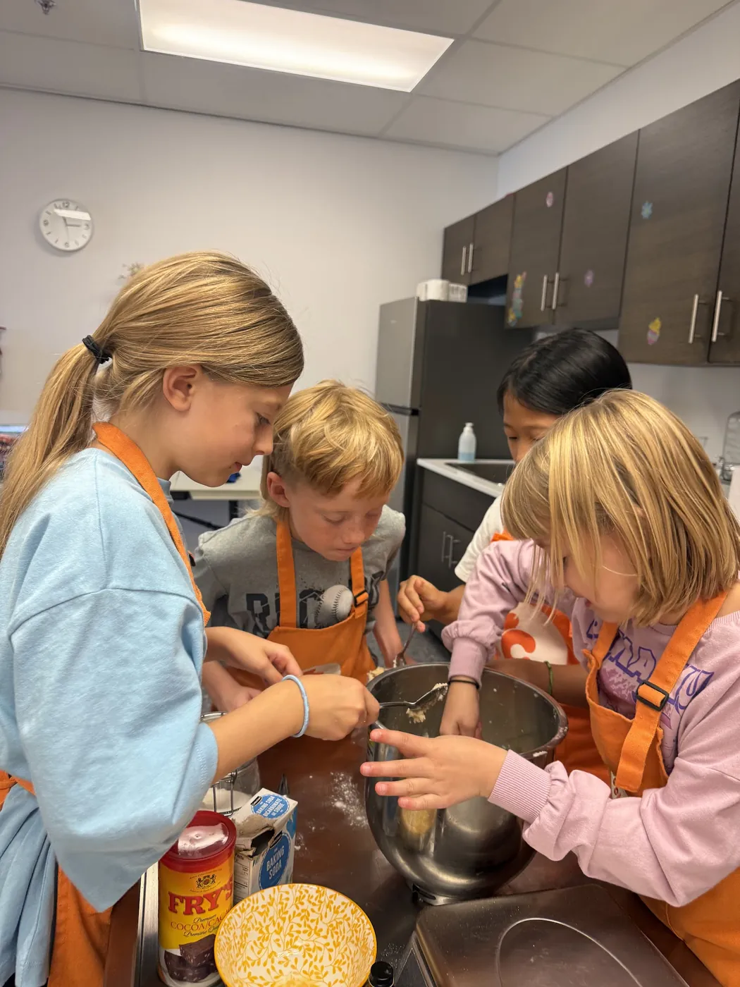 Four children in orange aprons mix dough in a large steel bowl during a Cookiddoo class
