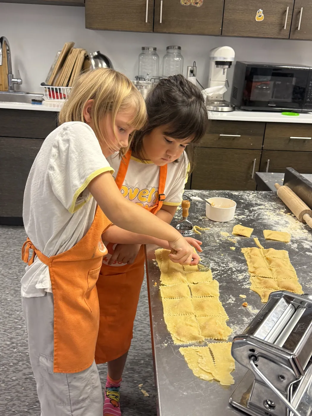 Kids feeding fresh pasta dough through a hand-crank pasta machine at a Cookiddoo cooking class
