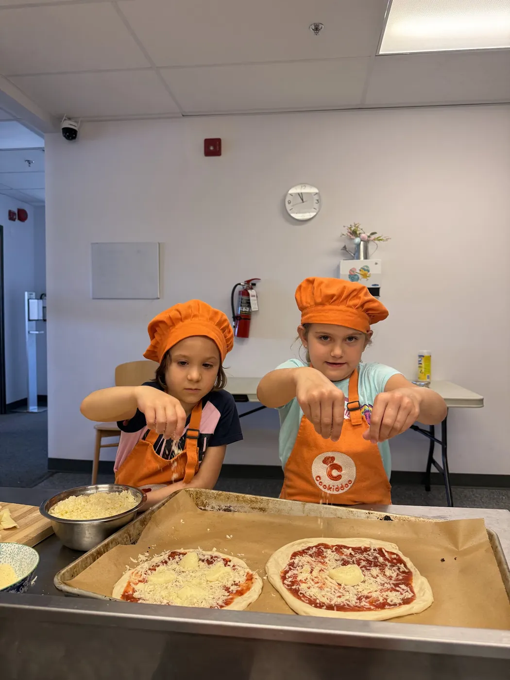 Two kids in Cookiddoo chef hats topping the pizzas they made from scratch at a cooking class
