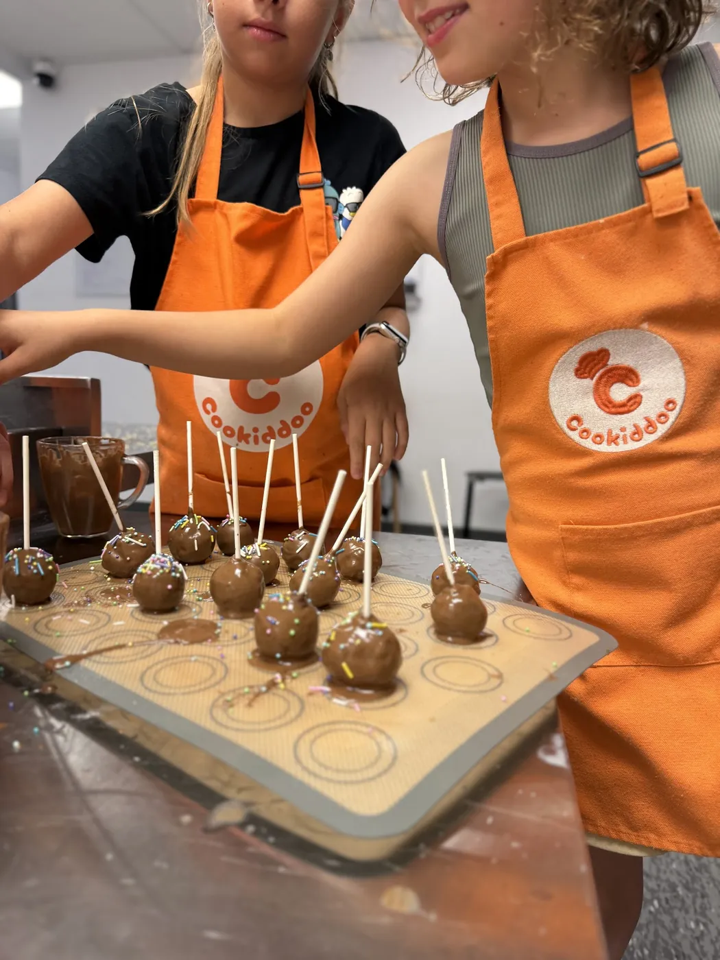 Deux enfants en tablier orange Cookiddoo déposent des cake pops au chocolat et aux vermicelles sur un tapis de cuisson