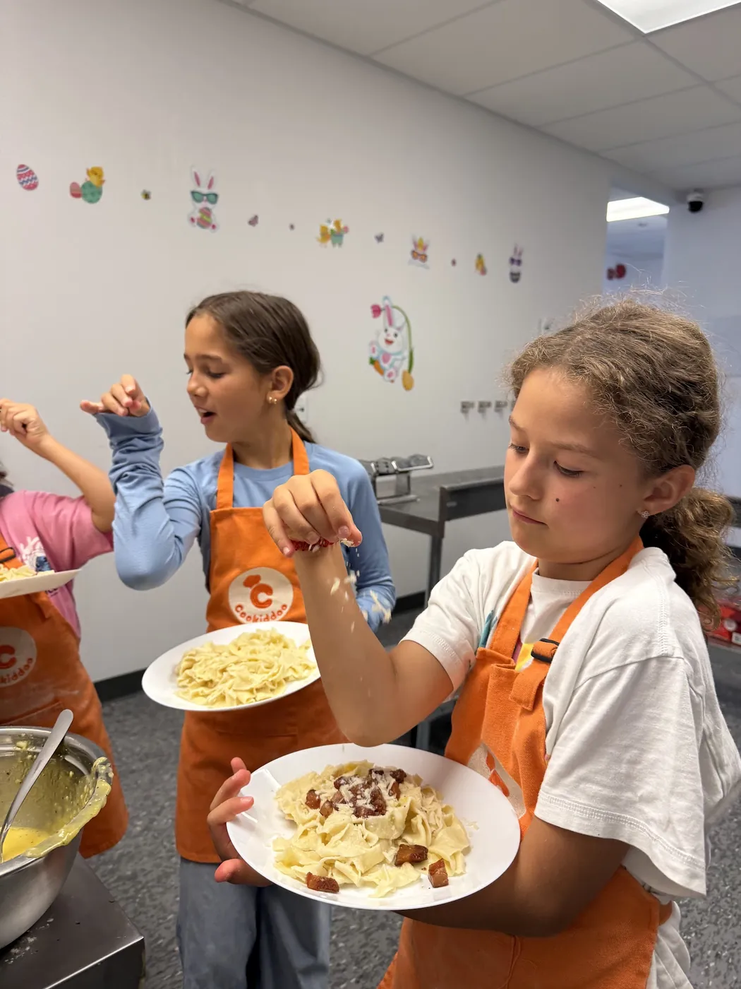 Une fillette en tablier orange Cookiddoo saupoudre de fromage des assiettes de pâtes carbonara