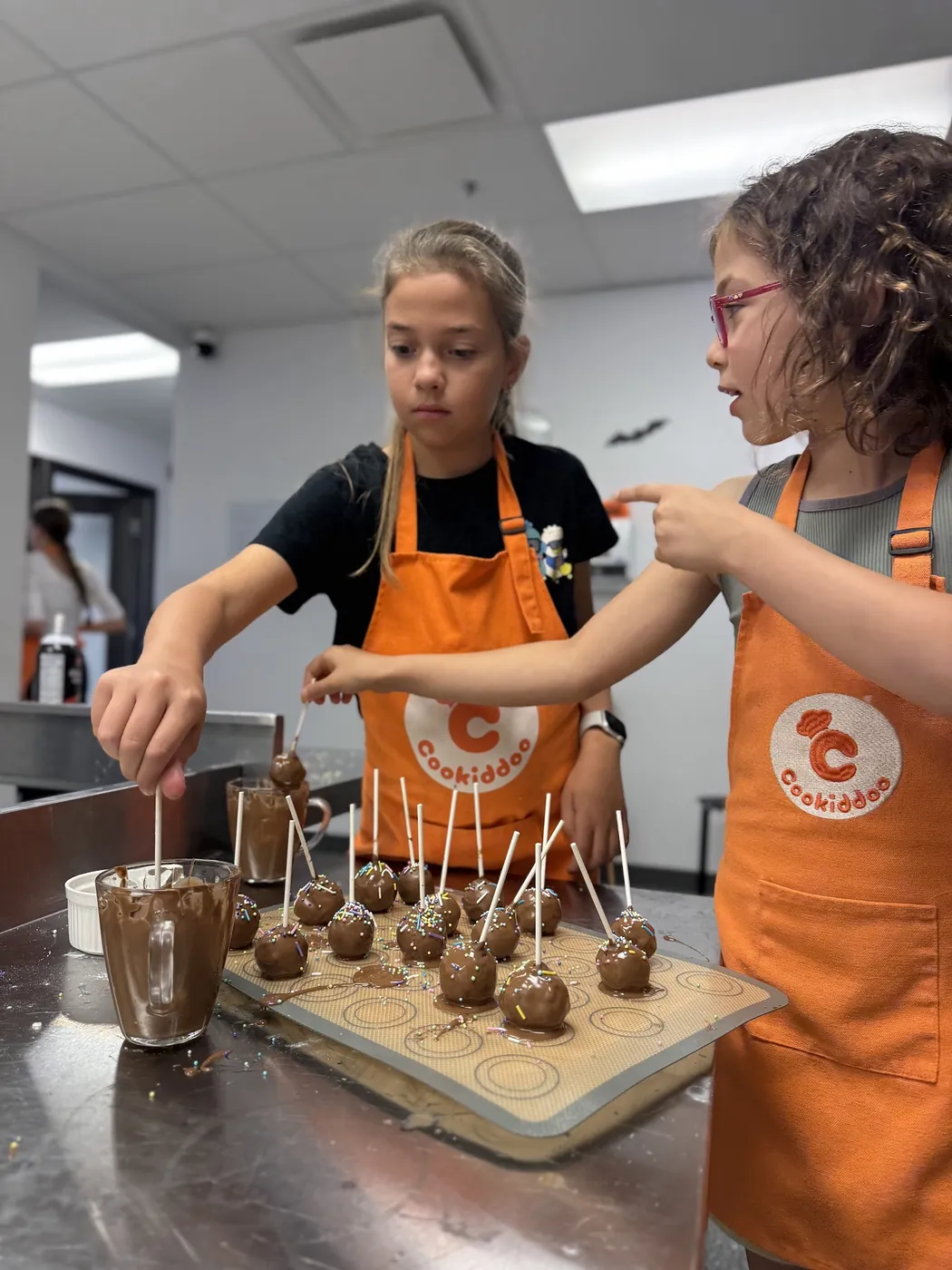 Deux filles en tablier orange Cookiddoo trempent des cake pops dans le chocolat et ajoutent des vermicelles