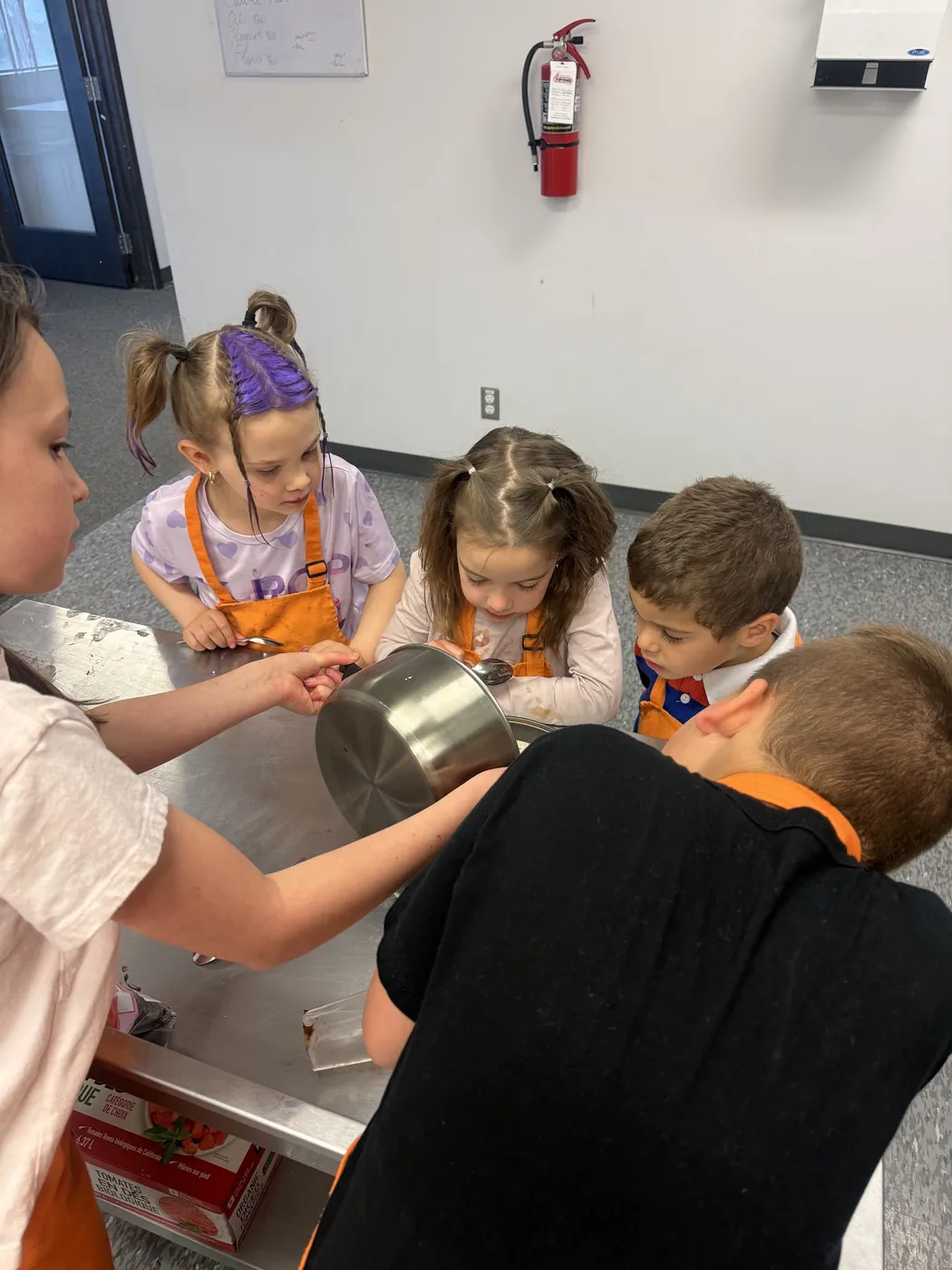 De jeunes enfants en tablier orange se penchent pendant qu’on verse une casserole sur un plan de travail en inox, lors d’un atelier de cuisine