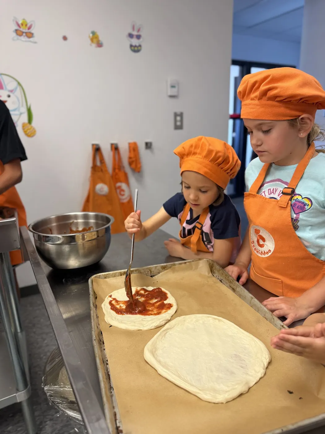 Enfants en toque orange Cookiddoo qui &eacute;talent la sauce sur leur p&acirc;te &agrave; pizza
