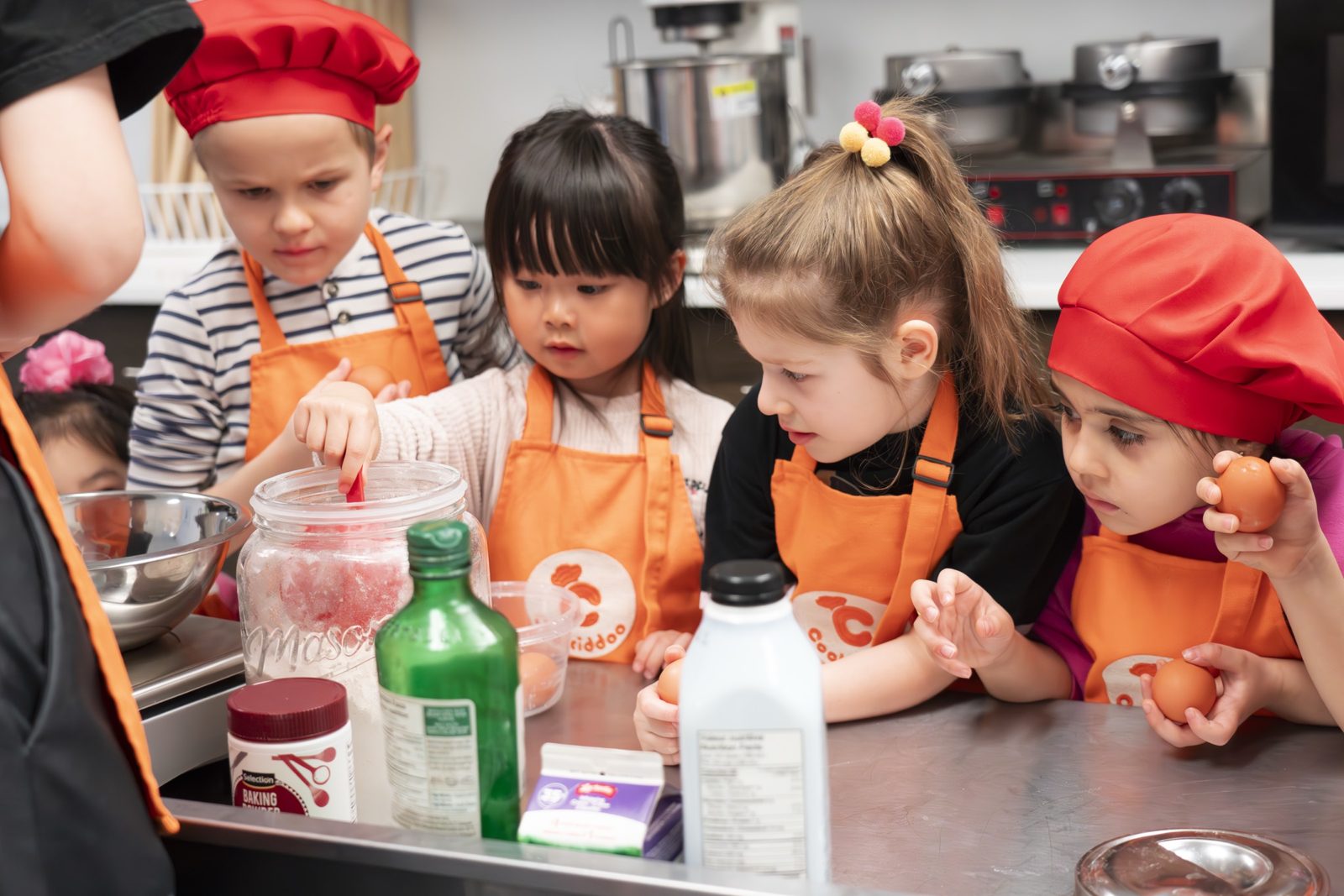 Діти у фартухах Cookiddoo розбивають яйця й відмірюють продукти біля сталевого столу